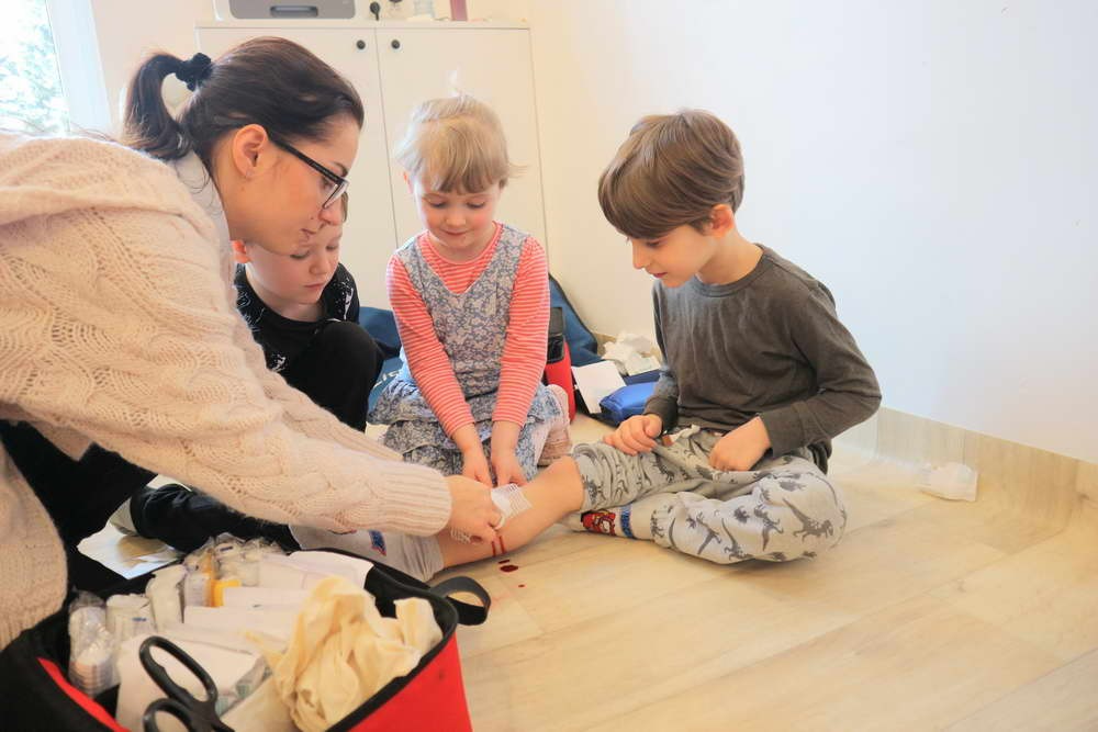 Kobieta w okularach opatruje ranę na ręce chłopca, obok siedzą dwoje dzieci, na podłodze rozłożona apteczka z wyposażeniem.