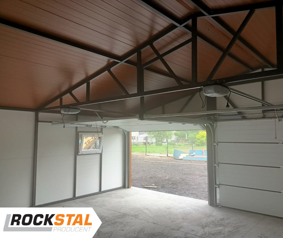Wnętrze garażu z bramą segmentową częściowo otwartą, widoczna konstrukcja stalowa dachu i okno w ścianie bocznej, logo producenta w lewym dolnym rogu.