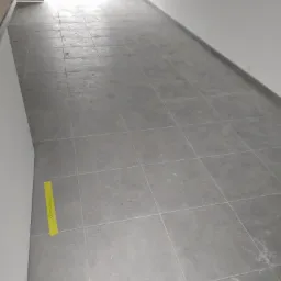 Szare płytki podłogowe ułożone w kwadraty, widoczna żółta taśma z napisem, surowy stan wnętrza przed wykończeniem, ściany pomalowane na biało, widoczne gniazdko elektryczne.