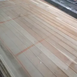 Jasne, drewniane deski podłogowe ułożone wzdłużnie, z widocznymi śladami pomarańczowej taśmy malarskiej, fragmentem ściany i elementami konstrukcyjnymi w tle.