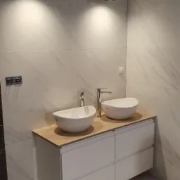 Nowoczesna łazienka z dwoma owalnymi umywalkami nablatowymi, drewnianym blatem i białymi szafkami podwieszanymi, oświetlona czarnymi lampami sufitowymi i wykończona marmurowymi płytkami ściennymi.