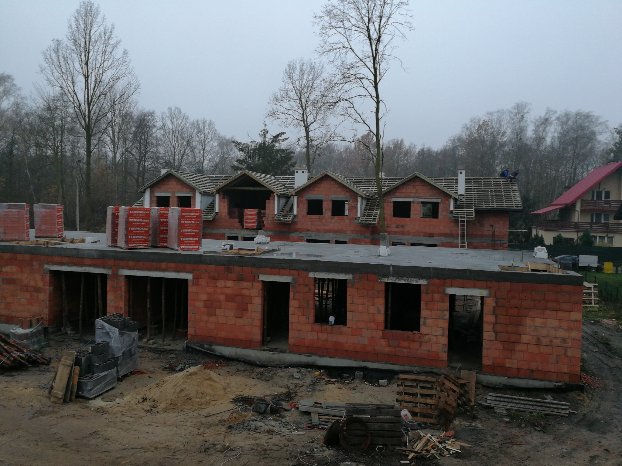 Surowy stan budowy szeregowców z czerwonej cegły, z widocznymi paletami cegieł na dachu jednego segmentu i dwoma pracownikami układającymi elementy więźby dachowej na innym segmencie, na tle...