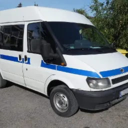 Biały Ford Transit z niebieskim pasem, zaparkowany na żwirowym podłożu, z widocznymi śmietnikami w tle.