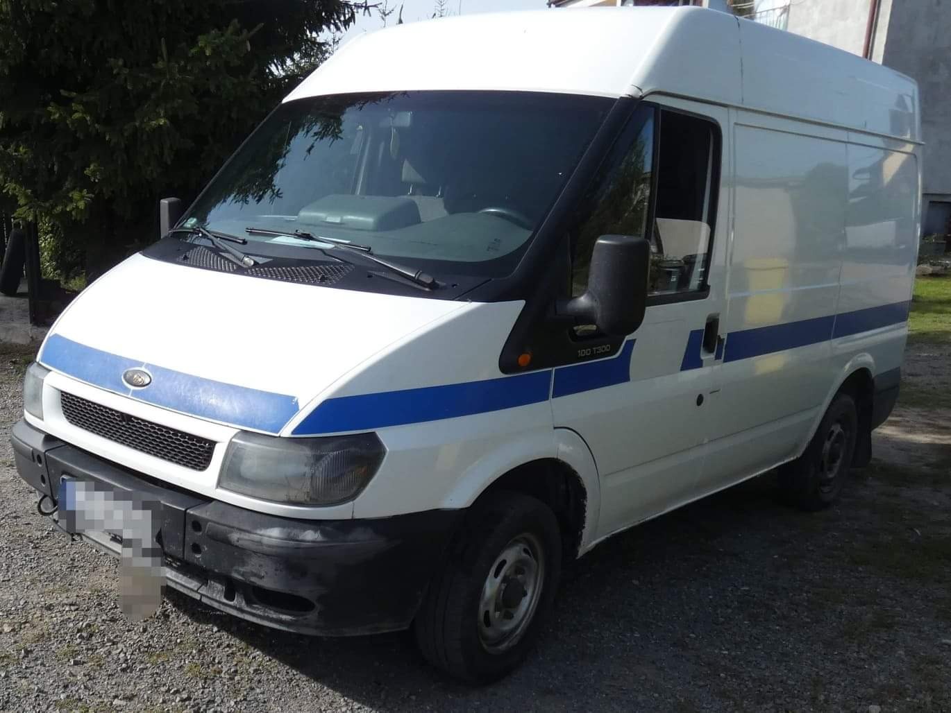 Biały Ford Transit z niebieskim pasem, zaparkowany na żwirowym podłożu, widoczny z perspektywy przedniego rogu.