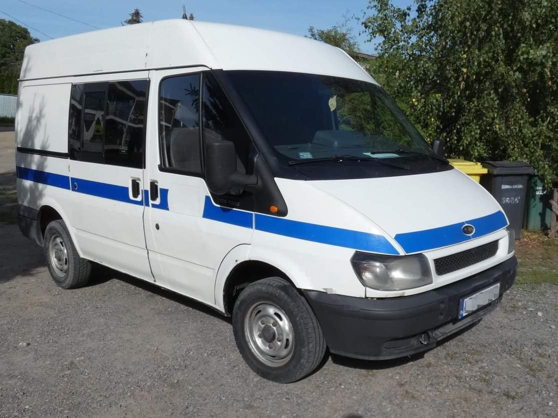 Biały Ford Transit z niebieskim pasem, zaparkowany na żwirowym podłożu, z widocznymi śmietnikami w tle.