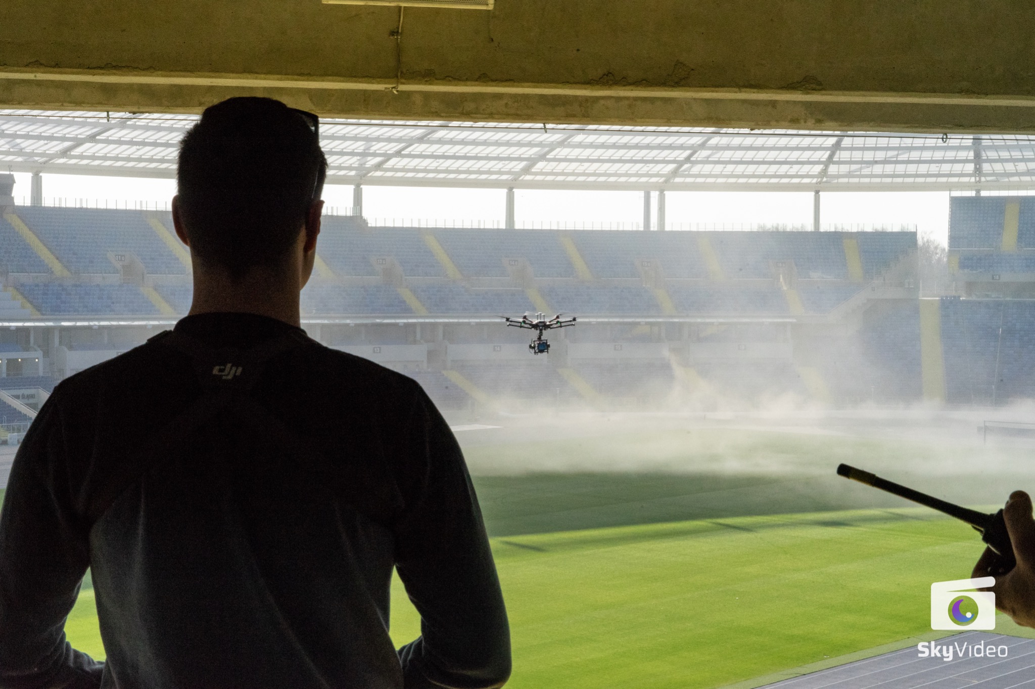 Operator drona obserwuje lot drona nad nawadnianym boiskiem stadionu, ujęcie zza pleców, widoczne logo DJI na koszulce operatora.