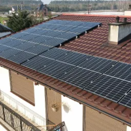 Panele słoneczne na brązowym dachu domu jednorodzinnego, widok z góry na balkon z drewnianą szpulą i resztki śniegu na trawniku.