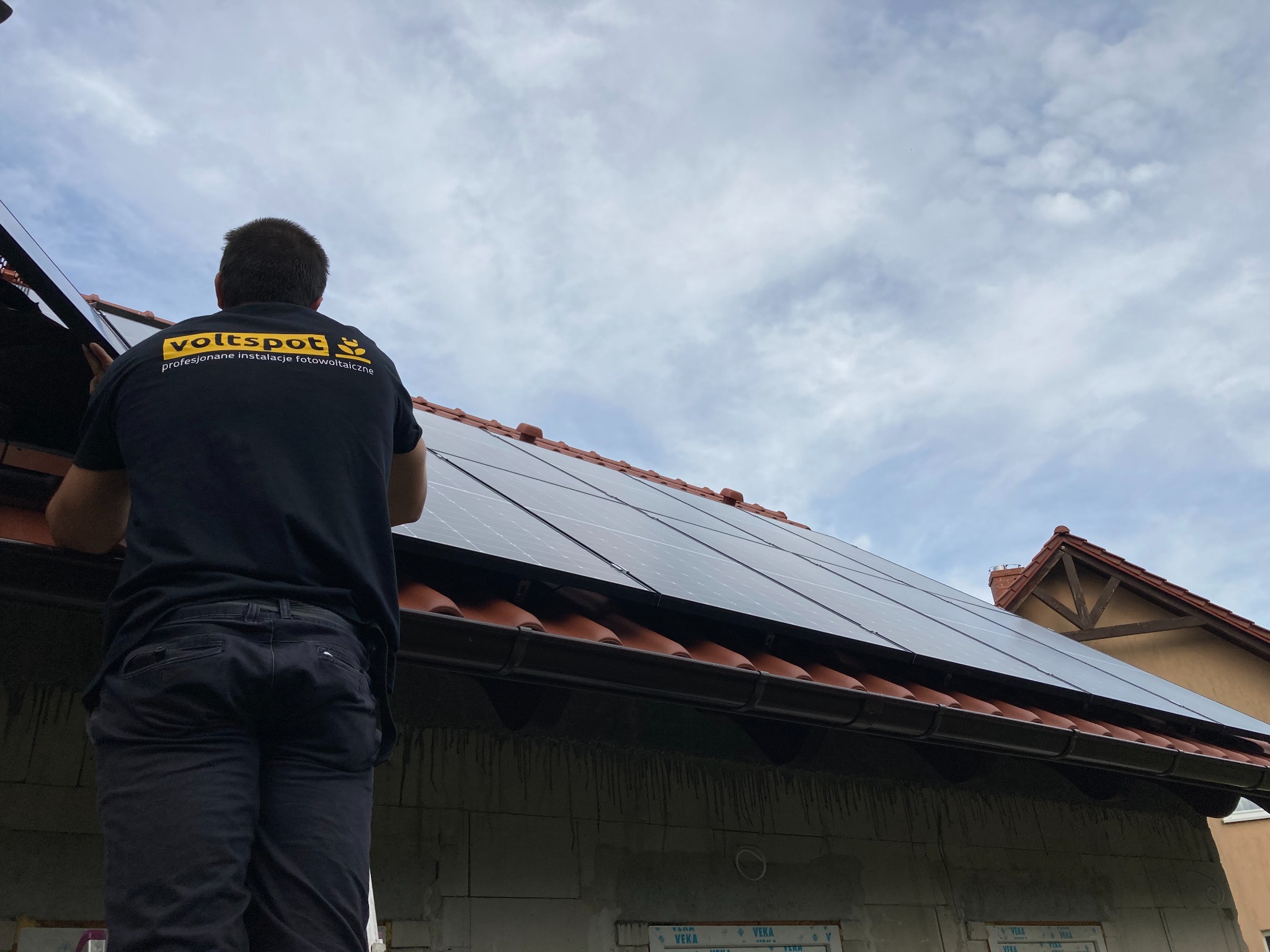 Instalator w czarnej koszulce z żółtym logo 'voltspot' montuje panele słoneczne na dachu domu z czerwoną dachówką, widok z dołu na tle pochmurnego nieba.