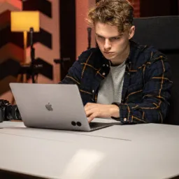 Młody mężczyzna z kręconymi włosami pracuje na laptopie, obok leży aparat fotograficzny z obiektywem, w tle ceglana ściana i lampa.