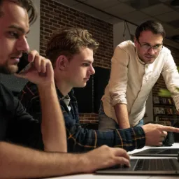 Trzech mężczyzn w biurze, jeden rozmawia przez telefon, drugi pisze na laptopie, trzeci wskazuje palcem na ekran laptopa, w tle ceglana ściana i roślina doniczkowa.