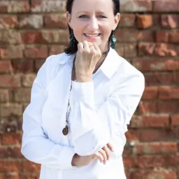 Portret uśmiechniętej kobiety w białej koszuli, z ręką pod brodą, na tle ceglanej ściany, z kolczykami z zielonymi kamieniami i naszyjnikiem z zawieszką w kształcie amonitu.