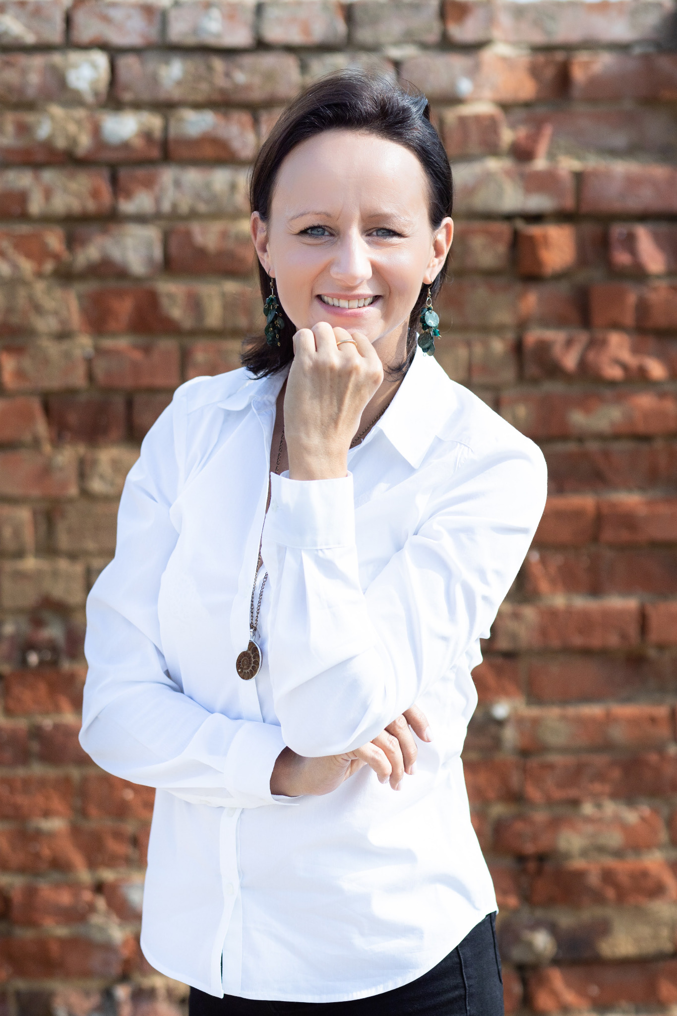 Portret uśmiechniętej kobiety w białej koszuli, z ręką pod brodą, na tle ceglanej ściany, z kolczykami z zielonymi kamieniami i naszyjnikiem z zawieszką w kształcie amonitu.