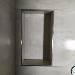 Wnęka prysznicowa wykończona płytkami w odcieniach beżu z metalową ramką, widoczny fragment instalacji z boku.