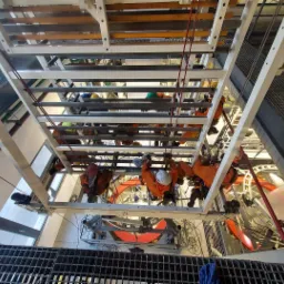 Widok z dołu na grupę pracowników w pomarańczowych kombinezonach, zabezpieczonych linami, pracujących na wysokości na konstrukcji stalowej wewnątrz budynku.