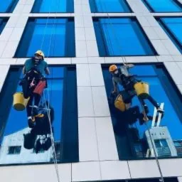 Dwóch pracowników wysokościowych myje okna biurowca na linach. Widoczne żółte wiadra, sprzęt alpinistyczny i odbicie sąsiednich budynków w szybach.