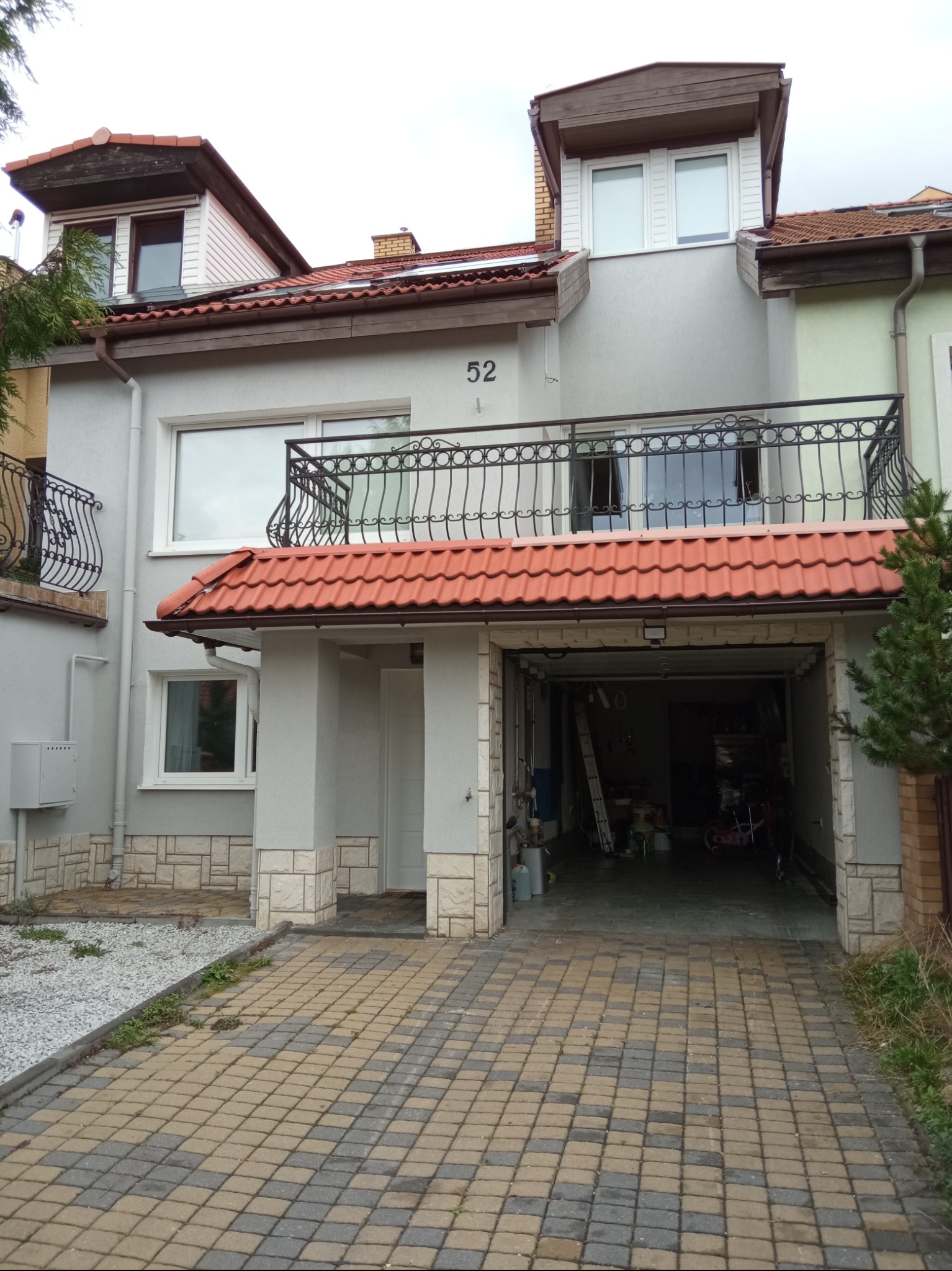 Elewacja szeregowego domu z garażem, numer 52, balkon z kutą balustradą, dachówka ceramiczna, podjazd z kostki brukowej, widoczne narzędzia i akcesoria w garażu.