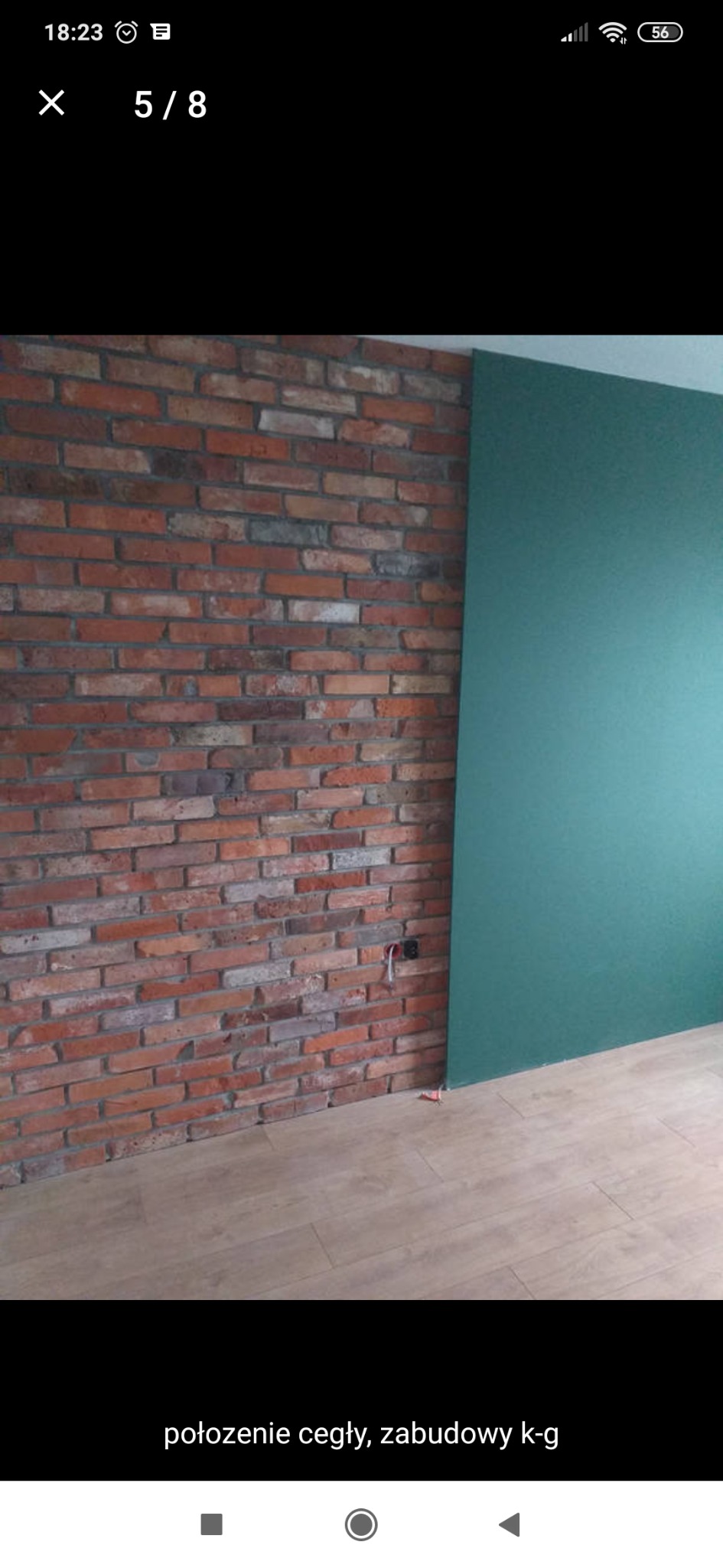 Ściana z czerwonej cegły sąsiadująca z gładką, zieloną ścianą, na podłodze panele podłogowe w odcieniach beżu, widoczne gniazdko elektryczne.