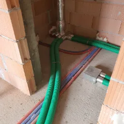 Wnętrze budynku w trakcie budowy z widocznymi przewodami wentylacyjnymi w kolorze zielonym, biegnącymi wzdłuż ściany z cegły dziurawki, oraz rurami w kolorach czerwonym i niebieskim, wychodzącymi...