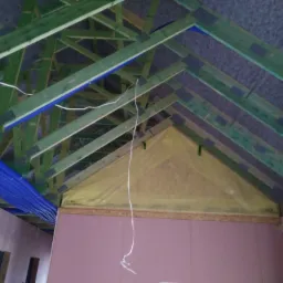 Wnętrze poddasza w trakcie adaptacji: widoczne ocieplenie, drewniana konstrukcja dachu pomalowana na zielono, niebieskie rury wentylacyjne oraz przewody elektryczne.