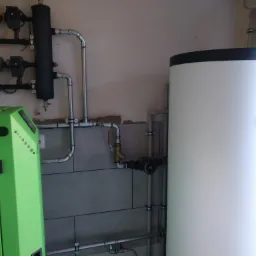 Zielony piec, biały zasobnik Kospel i instalacja CO z pompami obiegowymi, manometrami i zaworami, na tle szarej ściany z płytek.