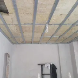 Poddasze w trakcie ocieplania wełną mineralną, widoczne metalowe profile sufitowe i fragment odsłoniętej ściany z cegły, drabina oparta o ścianę.