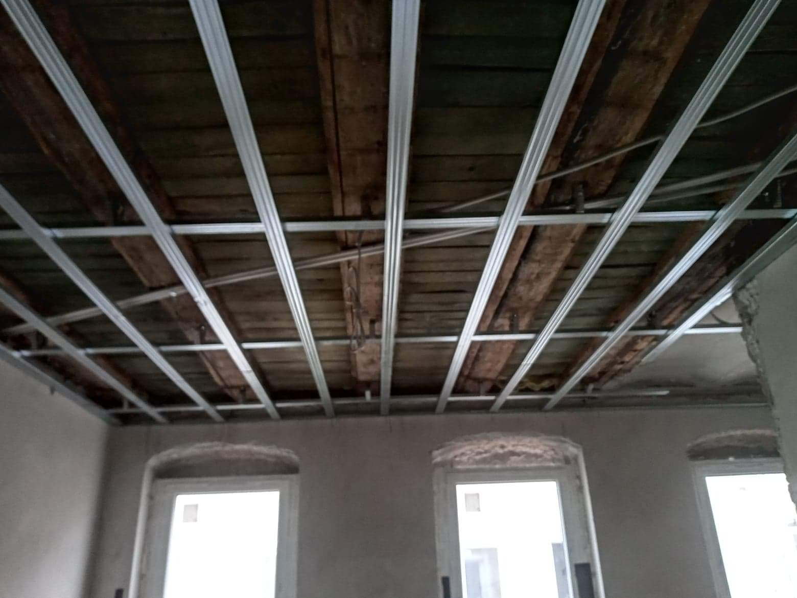Montaż metalowego stelaża sufitu podwieszanego na tle drewnianego stropu w pomieszczeniu z trzema oknami przed tynkowaniem ścian.