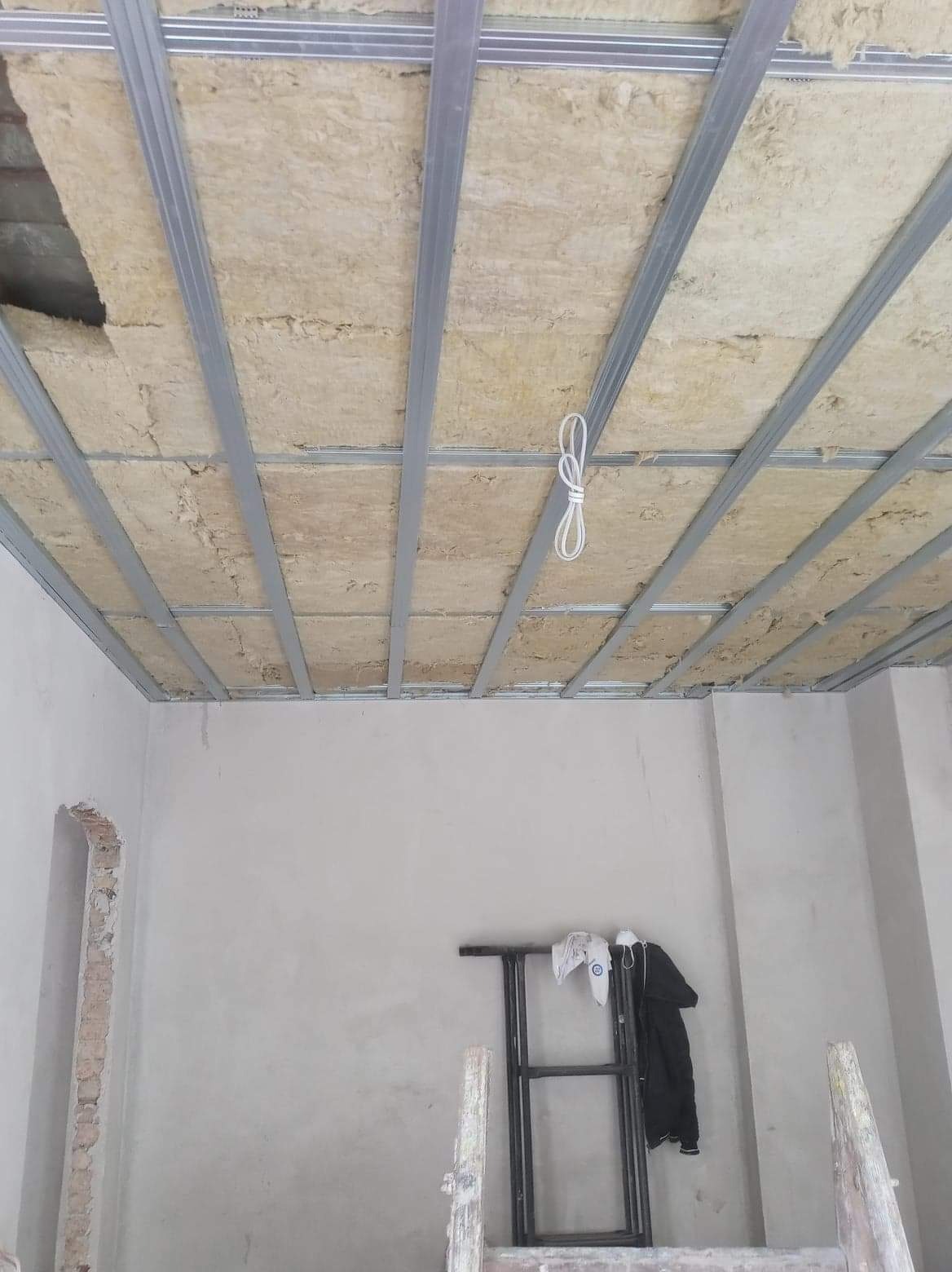 Poddasze w trakcie ocieplania wełną mineralną, widoczne metalowe profile sufitowe i fragment odsłoniętej ściany z cegły, drabina oparta o ścianę.