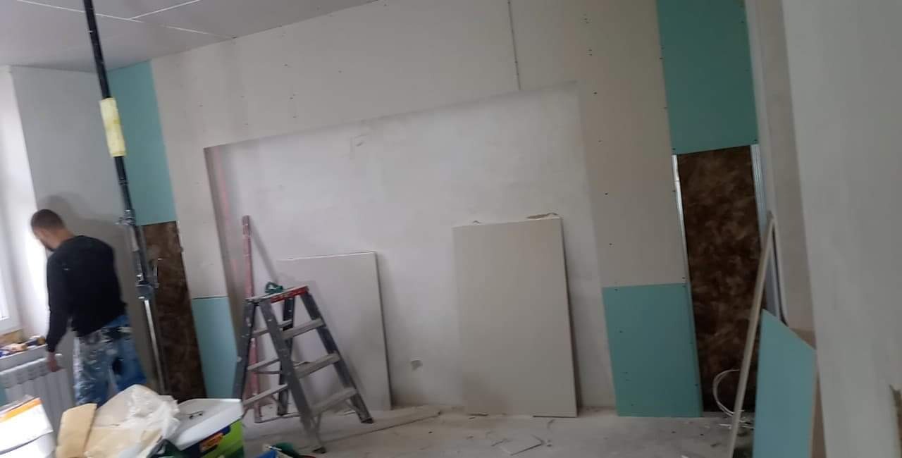 Wnętrze w trakcie remontu: płyty gipsowo-kartonowe zamontowane na ścianach, widoczna drabina i narzędzia, pracownik w tle przy oknie.