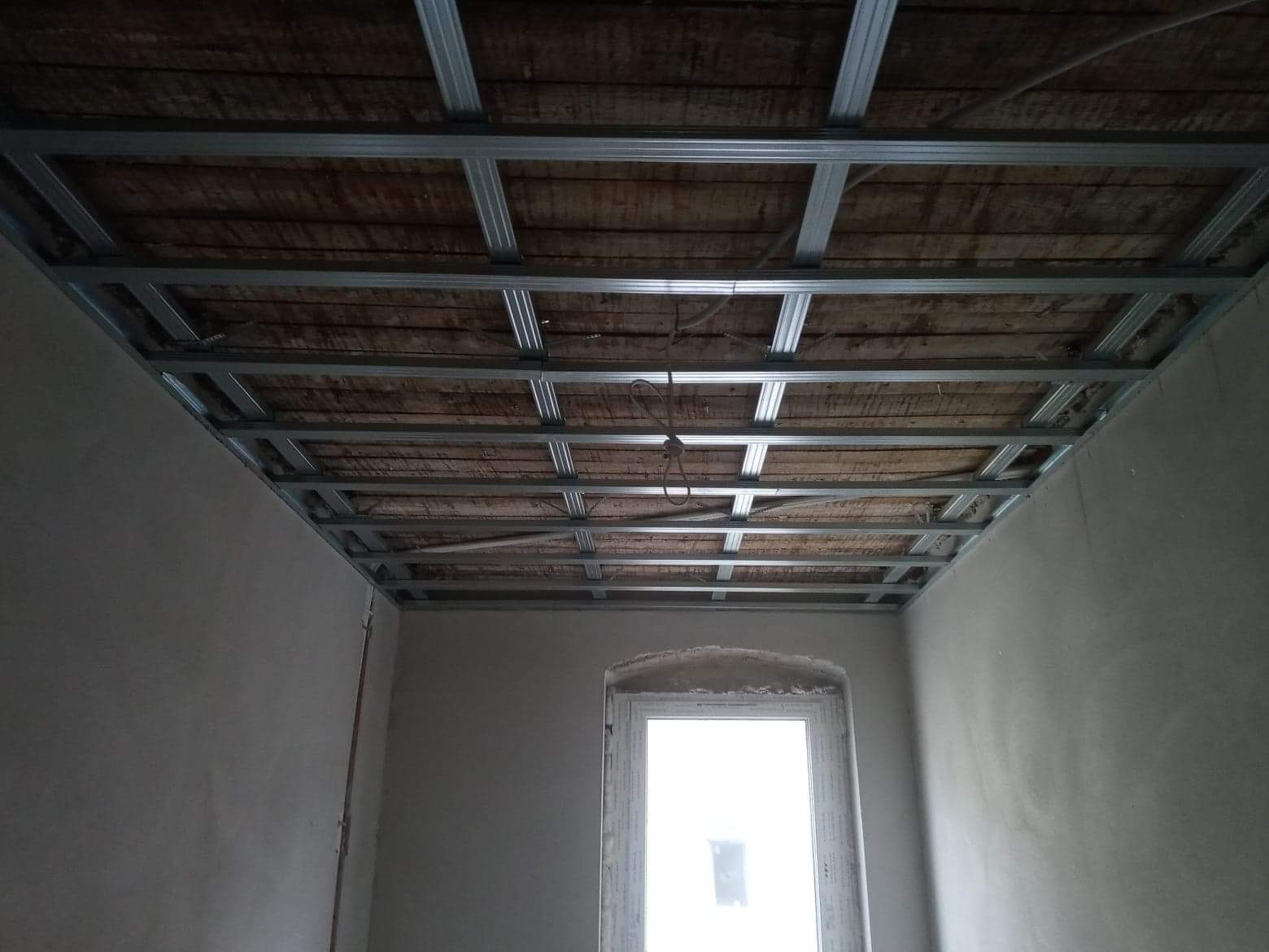 Stelaż z profili metalowych pod sufitem podwieszanym, widoczne drewniane deski stropu, surowe ściany w nowym budownictwie, okno z parapetem.