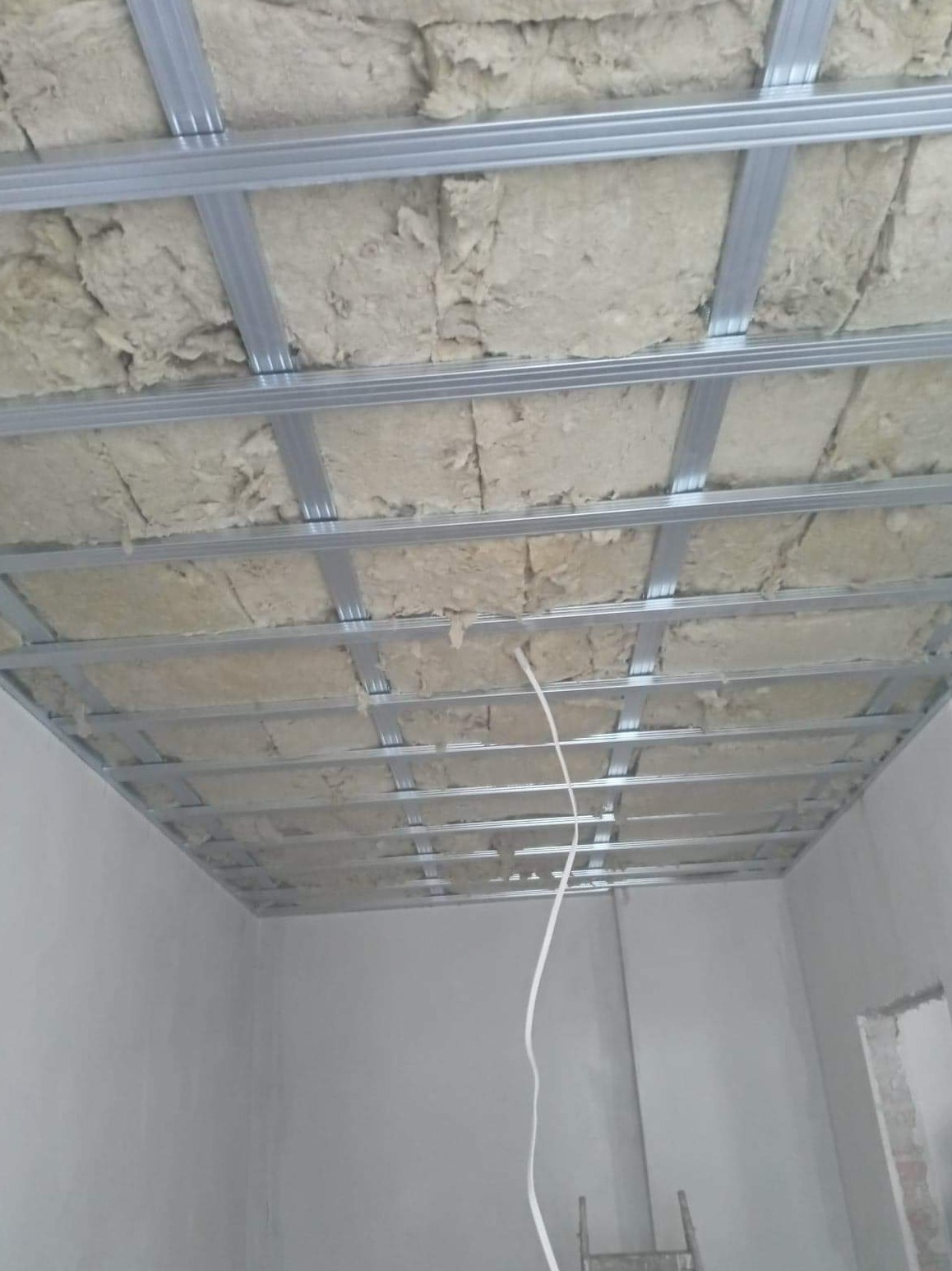 Montaż sufitu podwieszanego z izolacją z wełny mineralnej i metalową konstrukcją, widoczne przewody elektryczne.