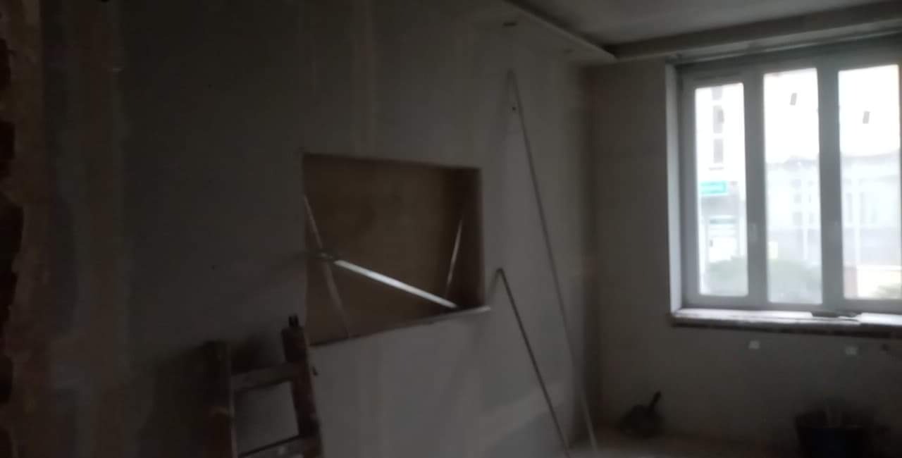 Wnętrze pokoju w trakcie remontu, ściany przygotowywane do wykończenia, widoczne szpachlowane fragmenty i drabina, okno z widokiem na ulicę.