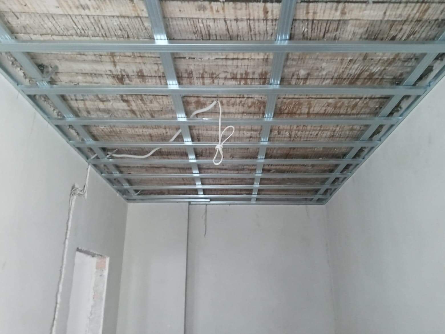 Stelaż metalowy sufitu podwieszanego zamontowany pod drewnianym stropem, widoczne przewody elektryczne i ściany w surowym stanie.
