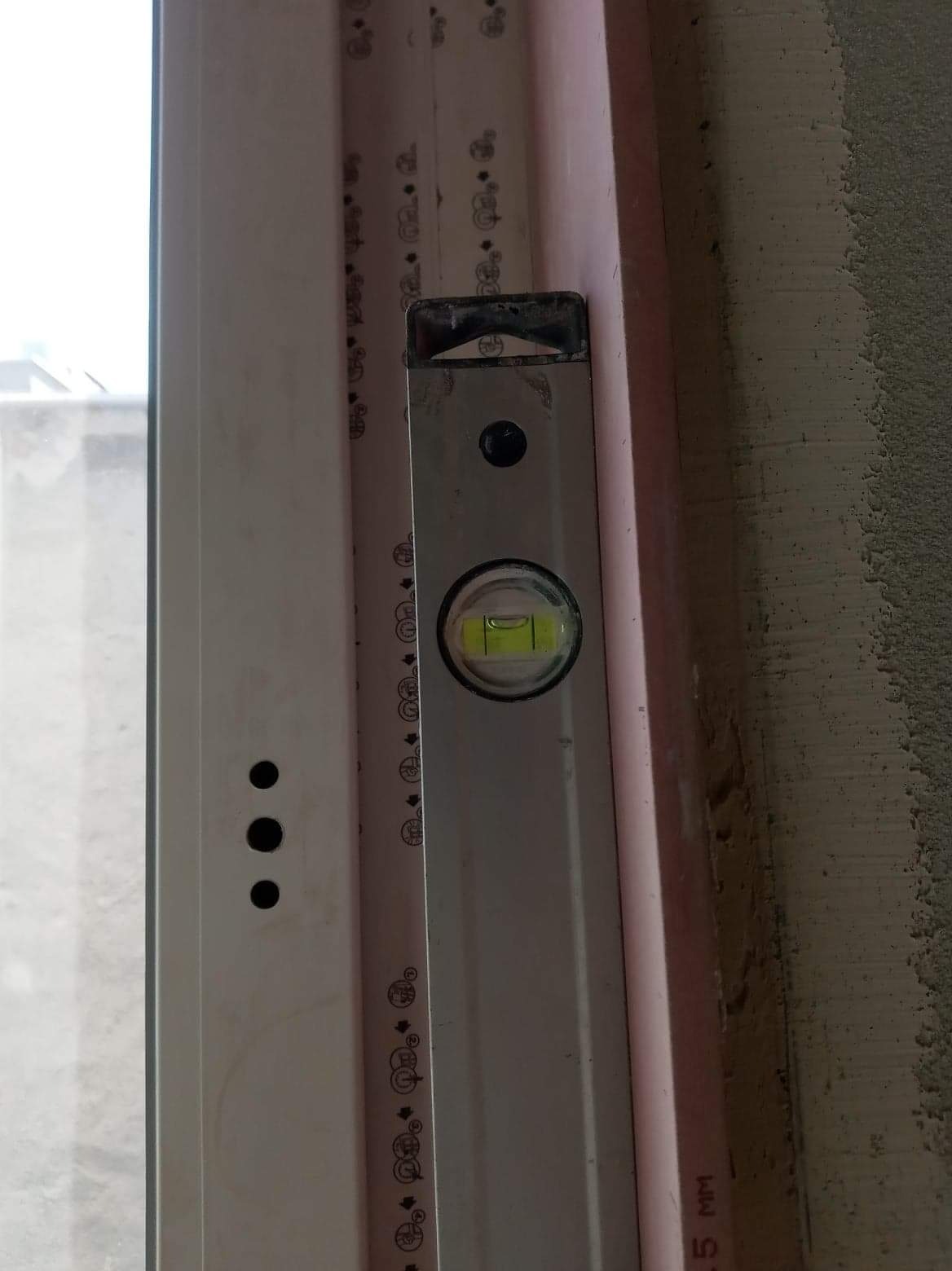 Poziomica przyłożona do ościeżnicy okiennej z widoczną pianką montażową i fragmentem surowej ściany.