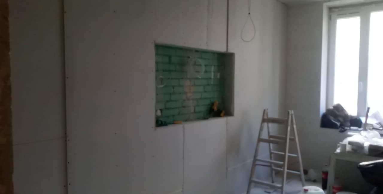 Wnętrze w trakcie prac wykończeniowych; ściany pokryte płytami gipsowo-kartonowymi, w ścianie wnęka z zieloną cegłą, drabina oparta o ścianę, widoczne kable wystające ze ściany.