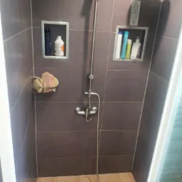 Kabina prysznicowa z szarymi płytkami, drewnopodobną podłogą, wnękami na kosmetyki i metalową armaturą, widok z dołu na głowicę prysznicową.