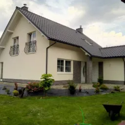 Jasna elewacja domu jednorodzinnego z ciemną dachówką i elementami dekoracyjnymi z kamienia, widok z ogrodu z fragmentem taczki.