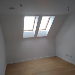 Poddasze z dwoma oknami dachowymi, drewnianą podłogą i białymi ścianami, widoczne gniazdka elektryczne i niezabezpieczone przewody oświetleniowe.