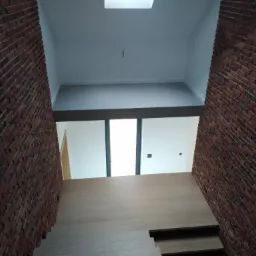 Klatka schodowa z drewnianymi schodami i ścianami wykończonymi czerwoną cegłą klinkierową, widok z dołu na górę, z widocznym oknem dachowym i fragmentem białej ściany na górze.