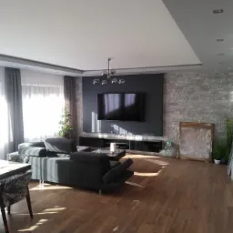 Jasne wnętrze salonu z szarą sofą, drewnianą podłogą i ścianą z kamienia dekoracyjnego, oświetlone przez duże okno z zasłonami.