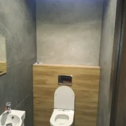 Nowoczesna toaleta podwieszana z drewnianą zabudową, umywalka narożna i szare płytki imitujące kamień, oświetlenie LED na suficie.