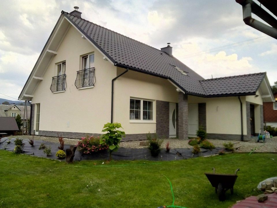 Jasna elewacja domu jednorodzinnego z ciemną dachówką i elementami dekoracyjnymi z kamienia, widok z ogrodu z fragmentem taczki.