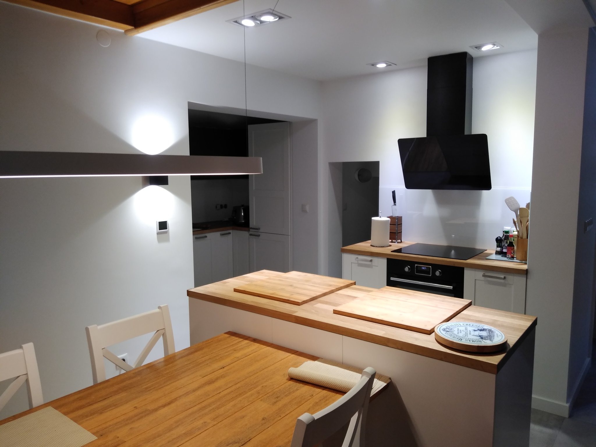 Nowoczesna kuchnia z drewnianym stołem, wyspą z drewnianym blatem, czarnym okapem i minimalistycznym oświetleniem.