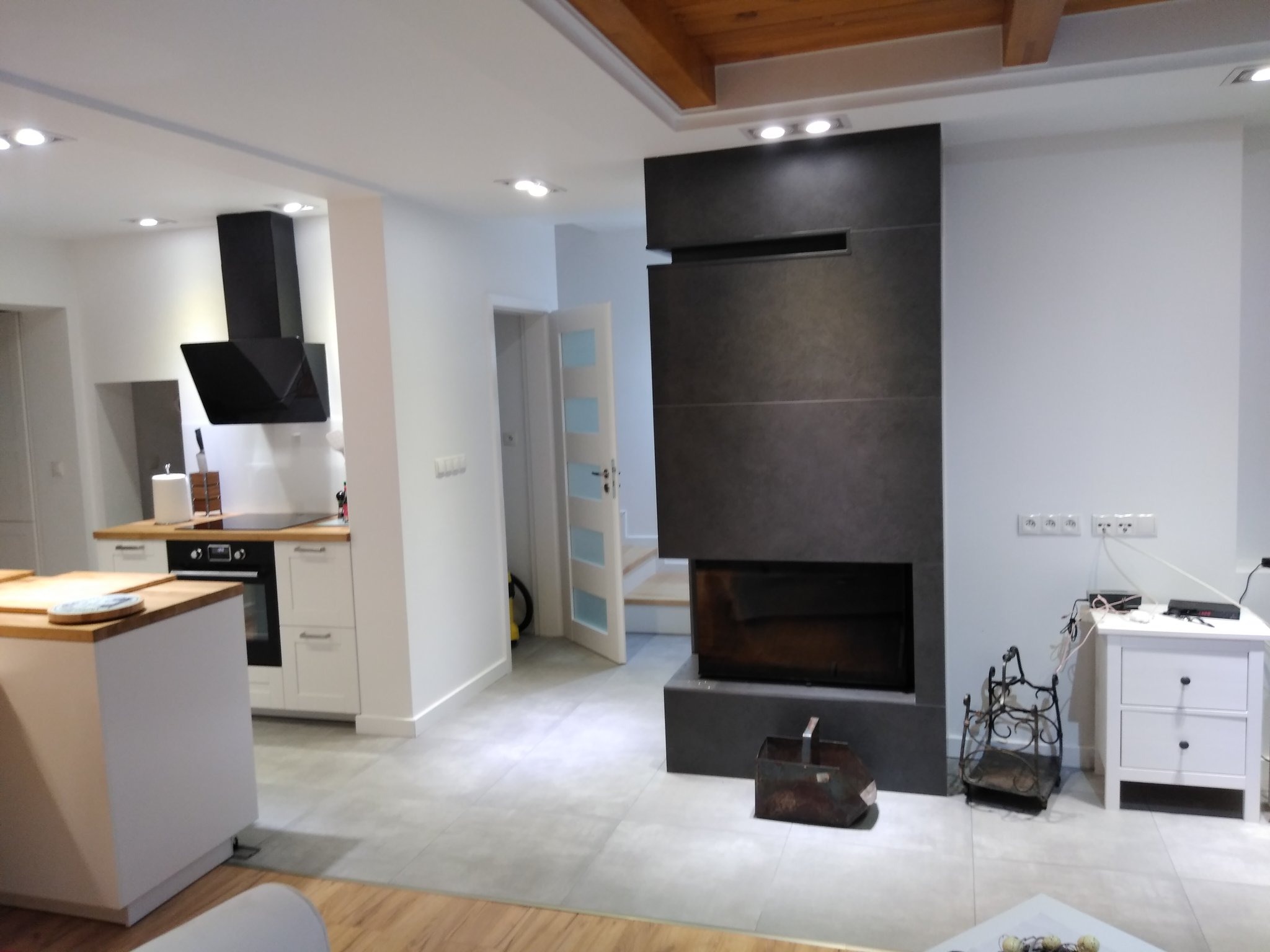 Nowoczesne wnętrze z szarą podłogą, białą kuchnią z drewnianym blatem, czarnym okapem i minimalistycznym kominkiem z ciemnego kamienia, drewniany sufit z belkami