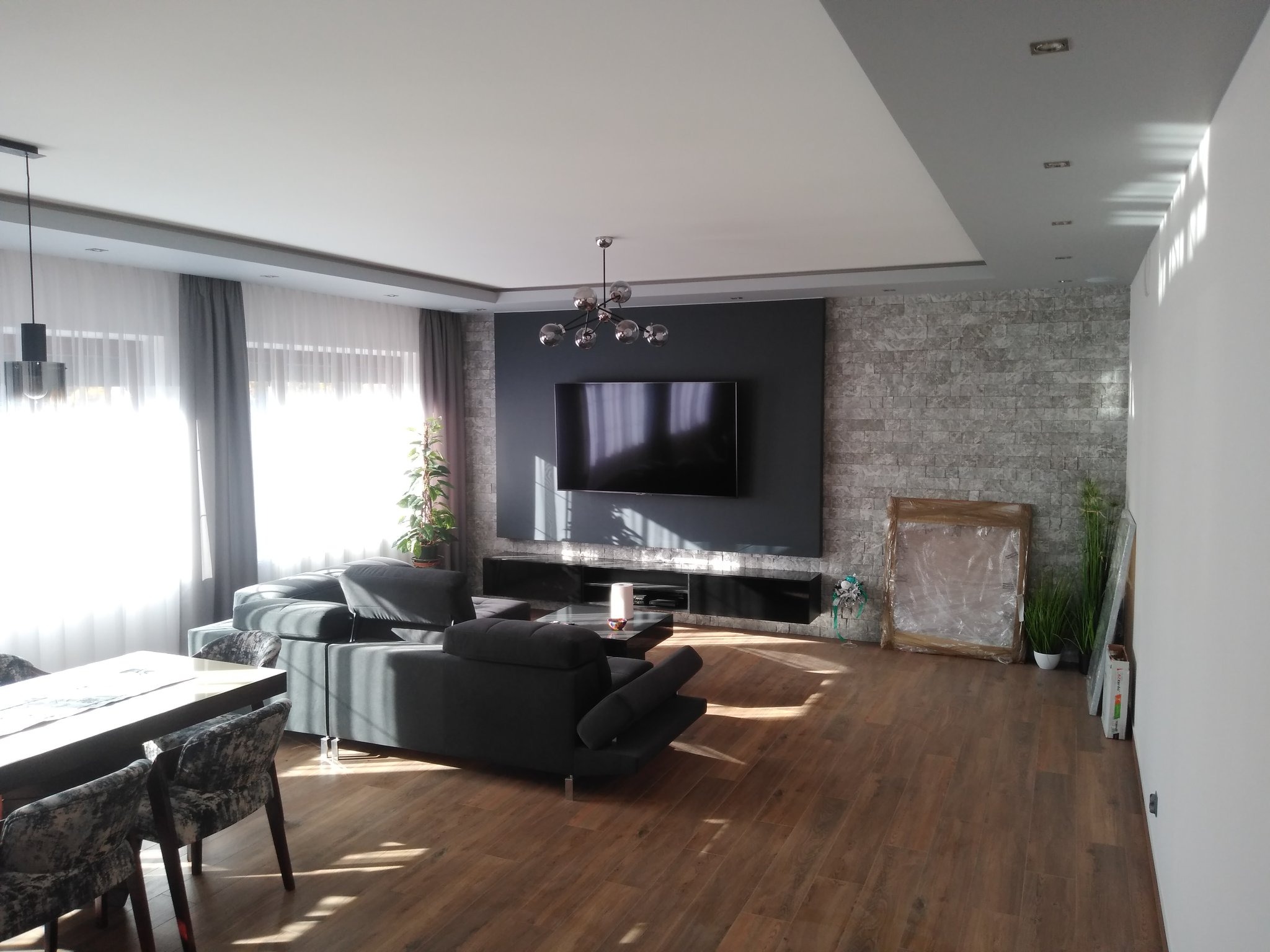 Jasne wnętrze salonu z szarą sofą, drewnianą podłogą i ścianą z kamienia dekoracyjnego, oświetlone przez duże okno z zasłonami.