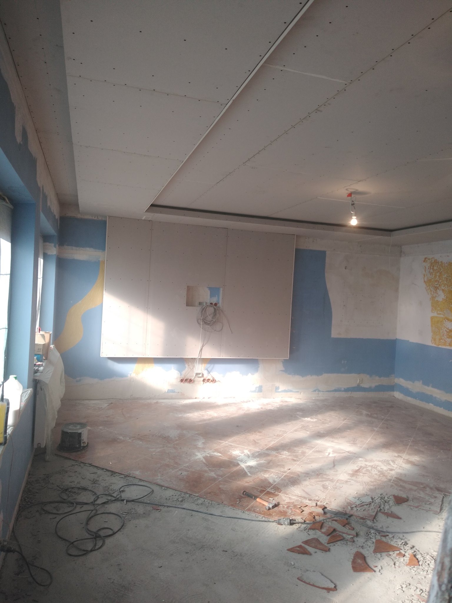 Wnętrze w trakcie remontu: częściowo zamontowana płyta gipsowo-kartonowa na ścianie z wystającymi przewodami, zdemontowana podłoga z widocznymi fragmentami płytek, ściany z resztkami starej farby...