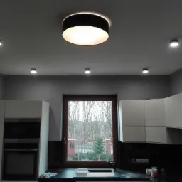 Nowoczesna kuchnia z białymi szafkami, czarnym blatem i dużym oknem z widokiem na drzewa, oświetlona okrągłą lampą sufitową i punktowymi reflektorami.