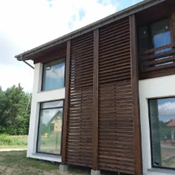 Nowoczesny dom z drewnianą pergolą w formie żaluzji, osłaniającą część fasady, widok z zewnątrz w słoneczny dzień.