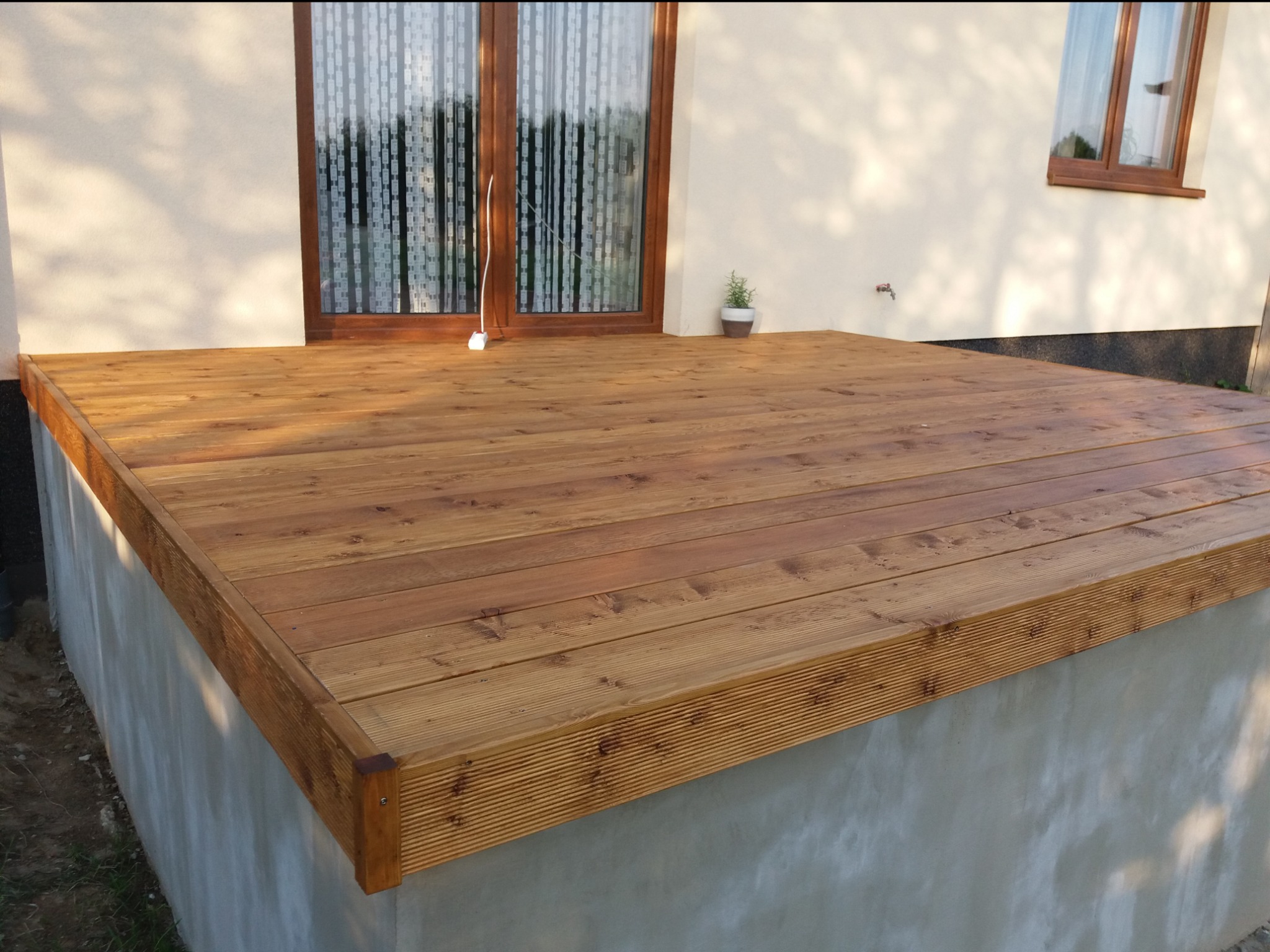 Nowoczesny taras z desek drewnianych, z subtelnym oświetleniem słonecznym i minimalistyczną dekoracją w postaci doniczki z ziołami przy oknie.