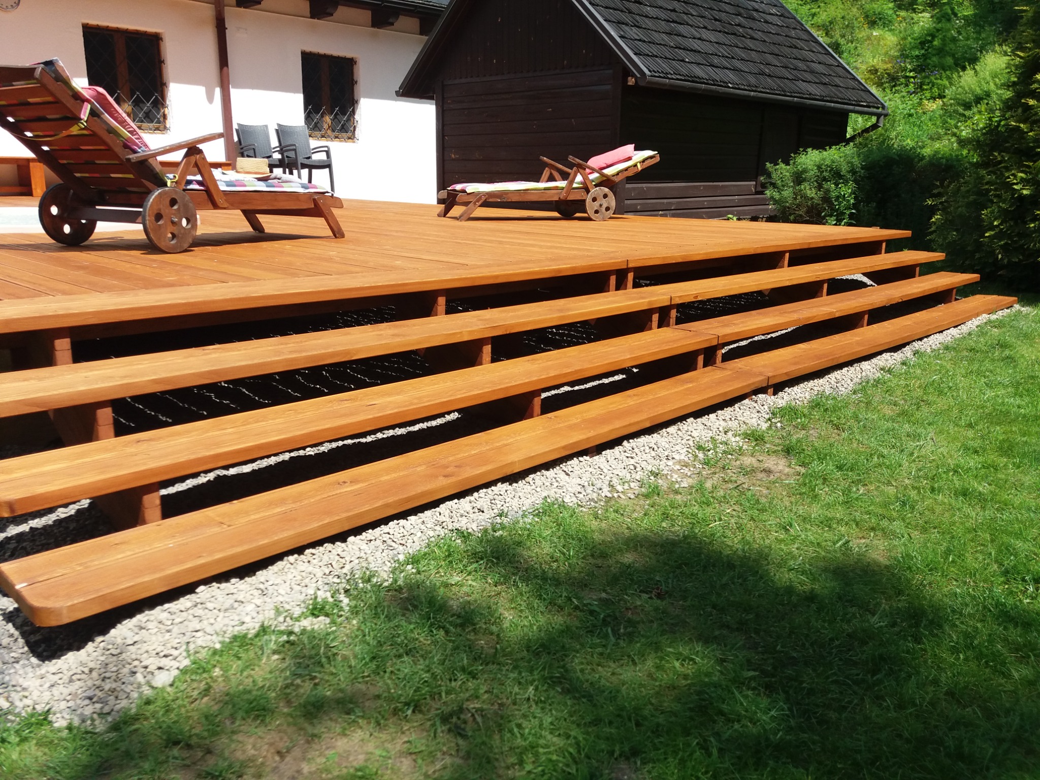 Nowoczesny, wielopoziomowy taras z drewna o ciepłym odcieniu, z leżakami na kółkach, otoczony zielonym trawnikiem i jasnymi kamykami.