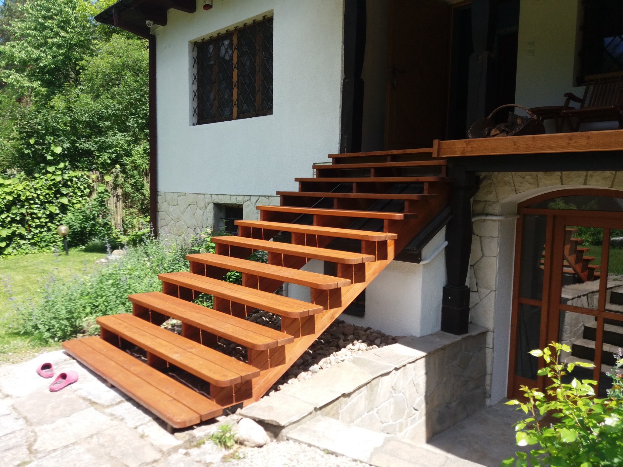 Zewnętrzne schody drewniane prowadzące na taras domu z białymi ścianami i kamienną podmurówką, widoczne różowe klapki na kamiennej posadzce.